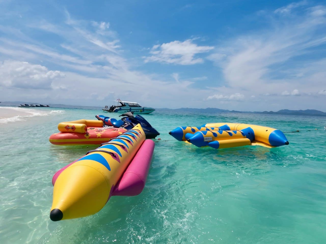 Yellow and blue inflatable banana boats floating beside a jet ski on clear turquoise water, with a speedboat in the distance