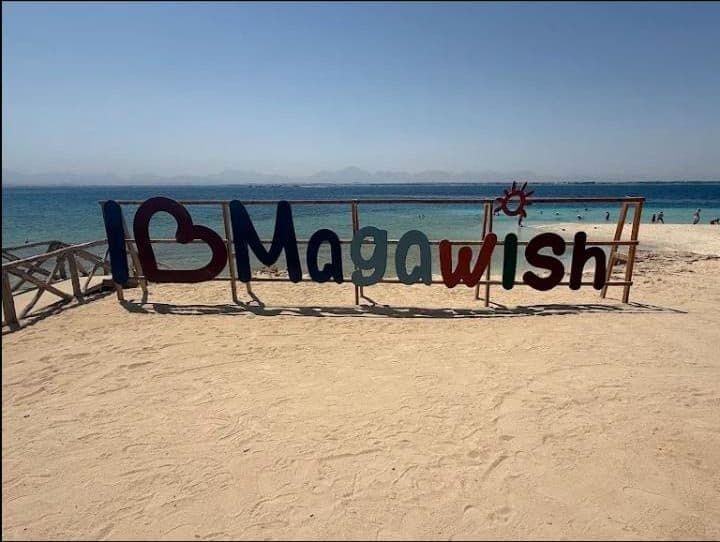 Colourful I Love Magawish sign on a sandy beach with calm turquoise sea and distant hills behind it