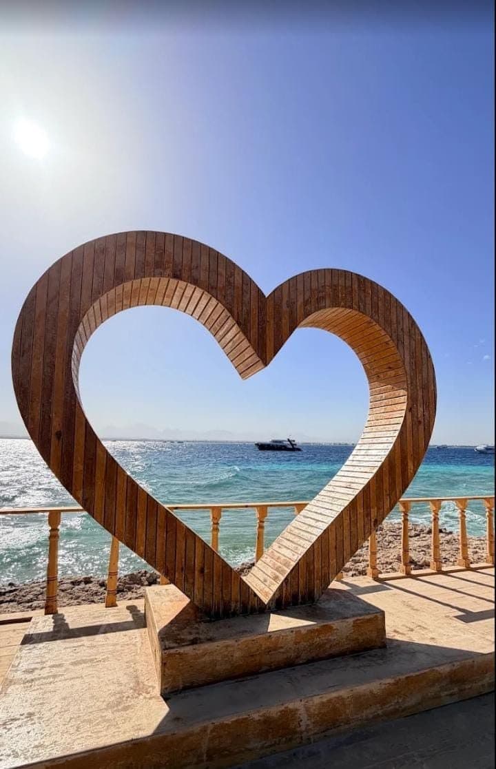Large wooden heart-shaped frame on a stone platform by the sea with a boat visible far out on the blue water