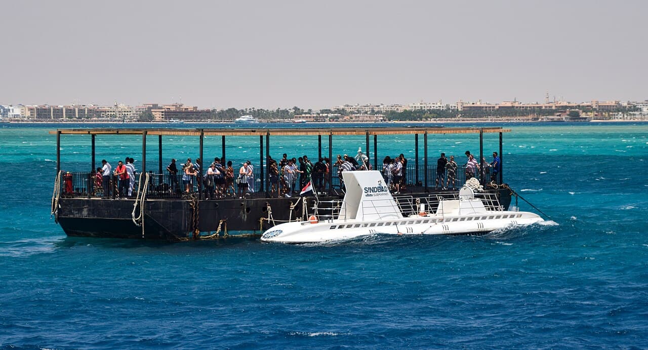 White Sindbad submarine moored beside a floating boarding platform where visitors wait, with the Hurghada shoreline behind